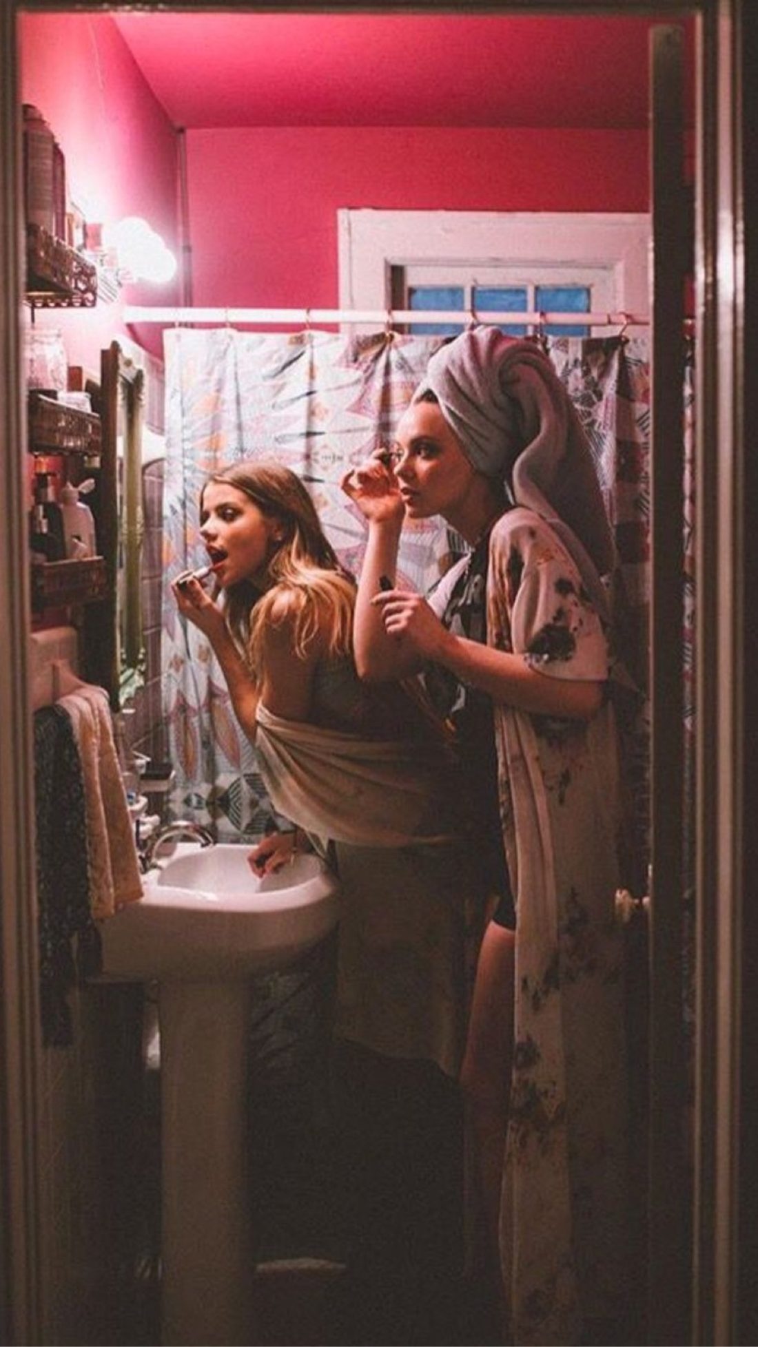 Two friends getting ready in a hot pink bathroom, seen through a doorway: one applying lipstick at a pedestal sink, the other in a robe and towel turban applying mascara